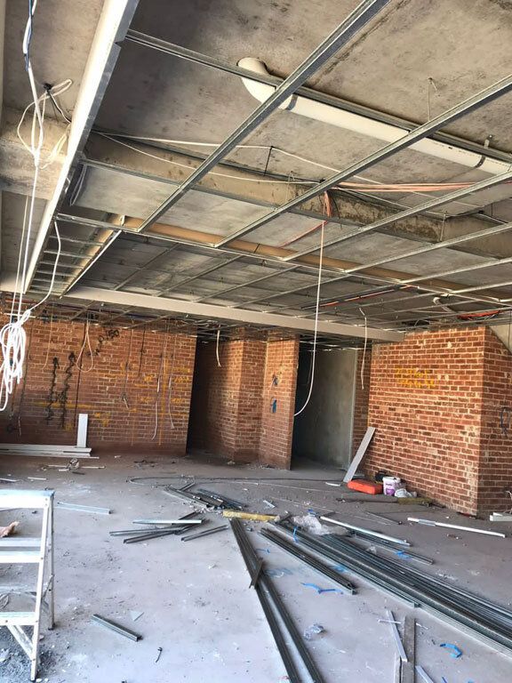 Steel furring channel hung on rods below a concrete slab in a stripped-out commercial space, conduit and light battens running above, brick walls and openings beyond
