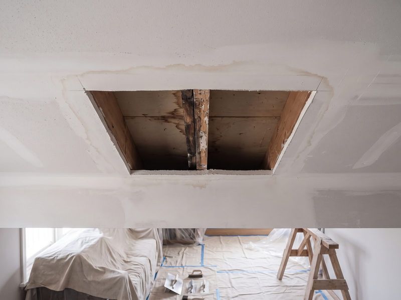 Water damaged section of ceiling plasterboard cut out and removed, exposing stained joists and a rotted batten above, with the tide-line feathering out across the surrounding ceiling