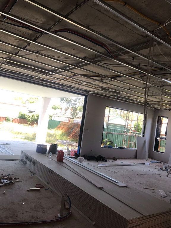 Ceiling furring and services running the full span of a wide commercial fitout before any plasterboard is fixed, with openings to the street beyond